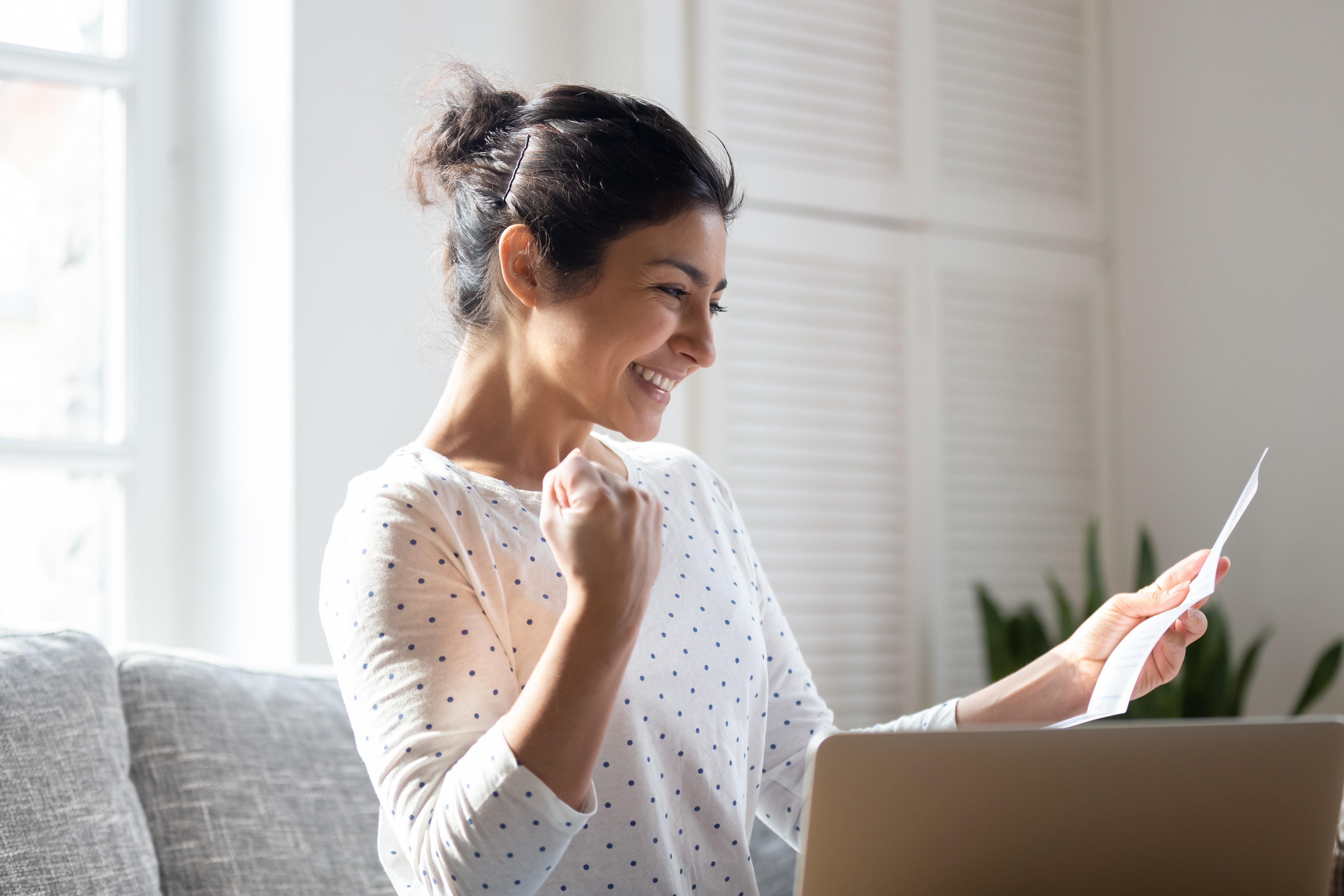 excited woman pumping fist and reading letter in sunlit living room